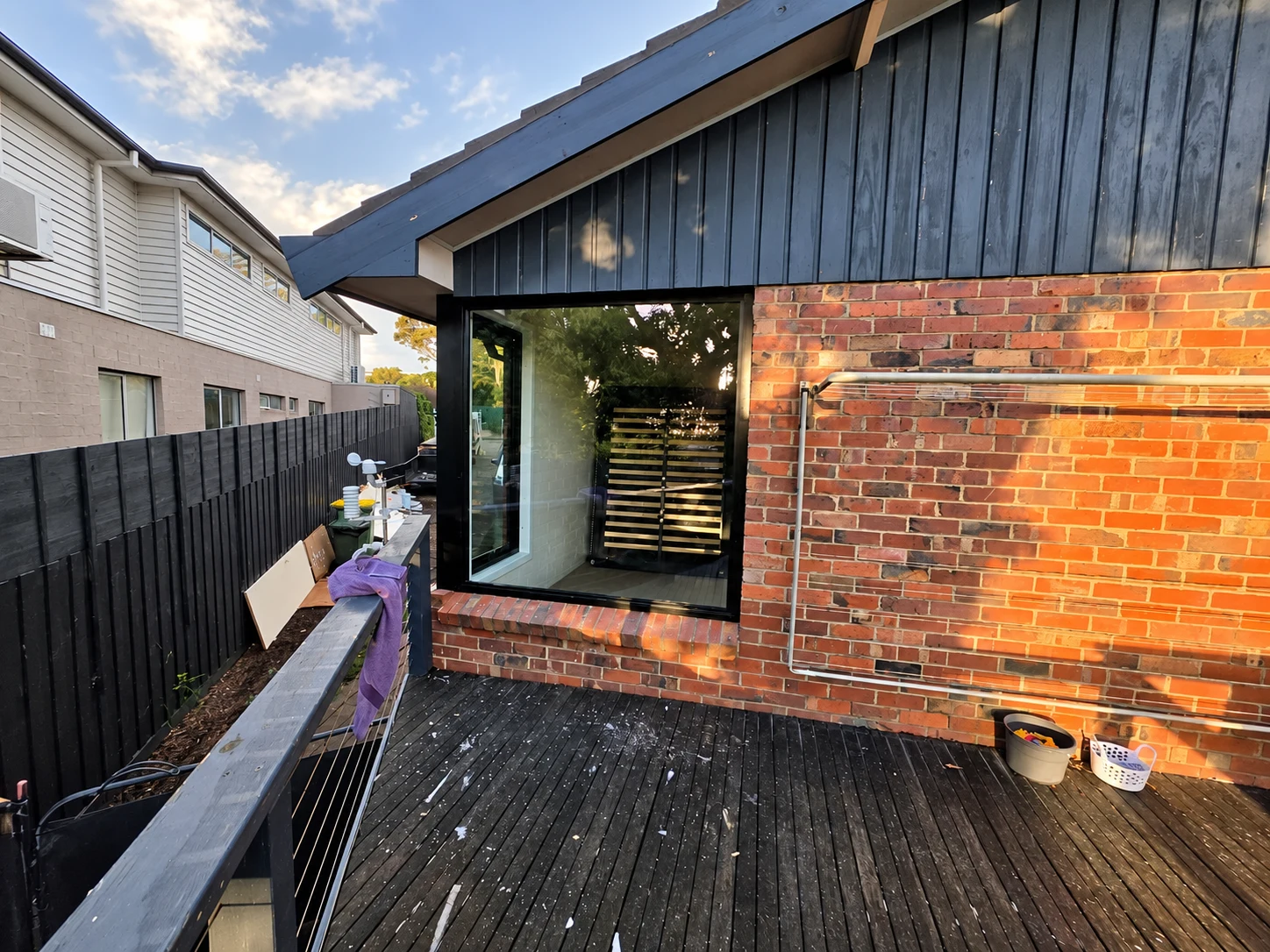 Black corner glazing beside a timber deck, red brick wall and dark weatherboard gable