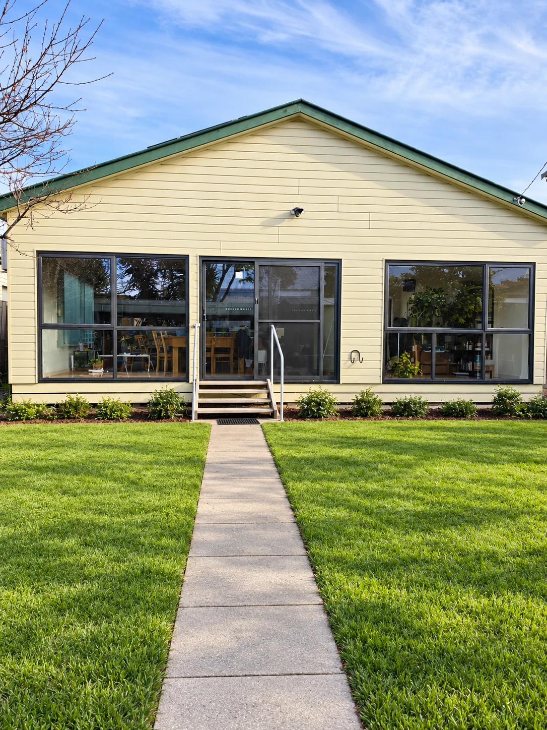 Pale yellow weatherboard home with dark aluminium windows, a central glazed door, neat green lawn and a clean garden path