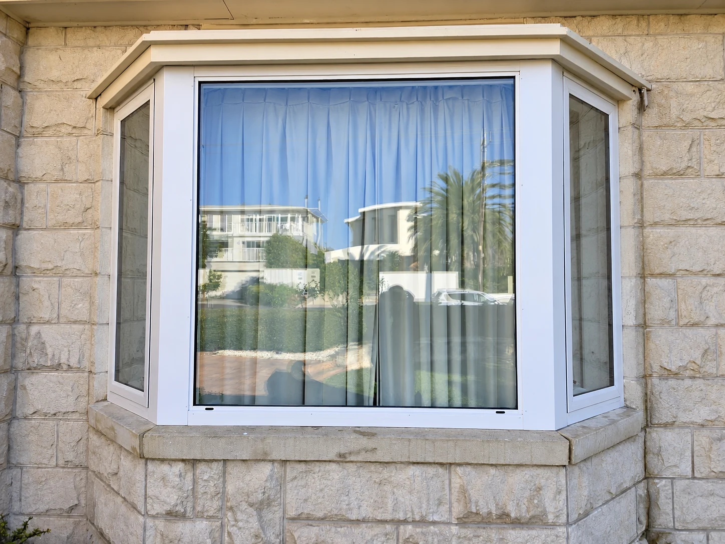 Close view of a white framed bay window in a stone wall with curtains behind the glass